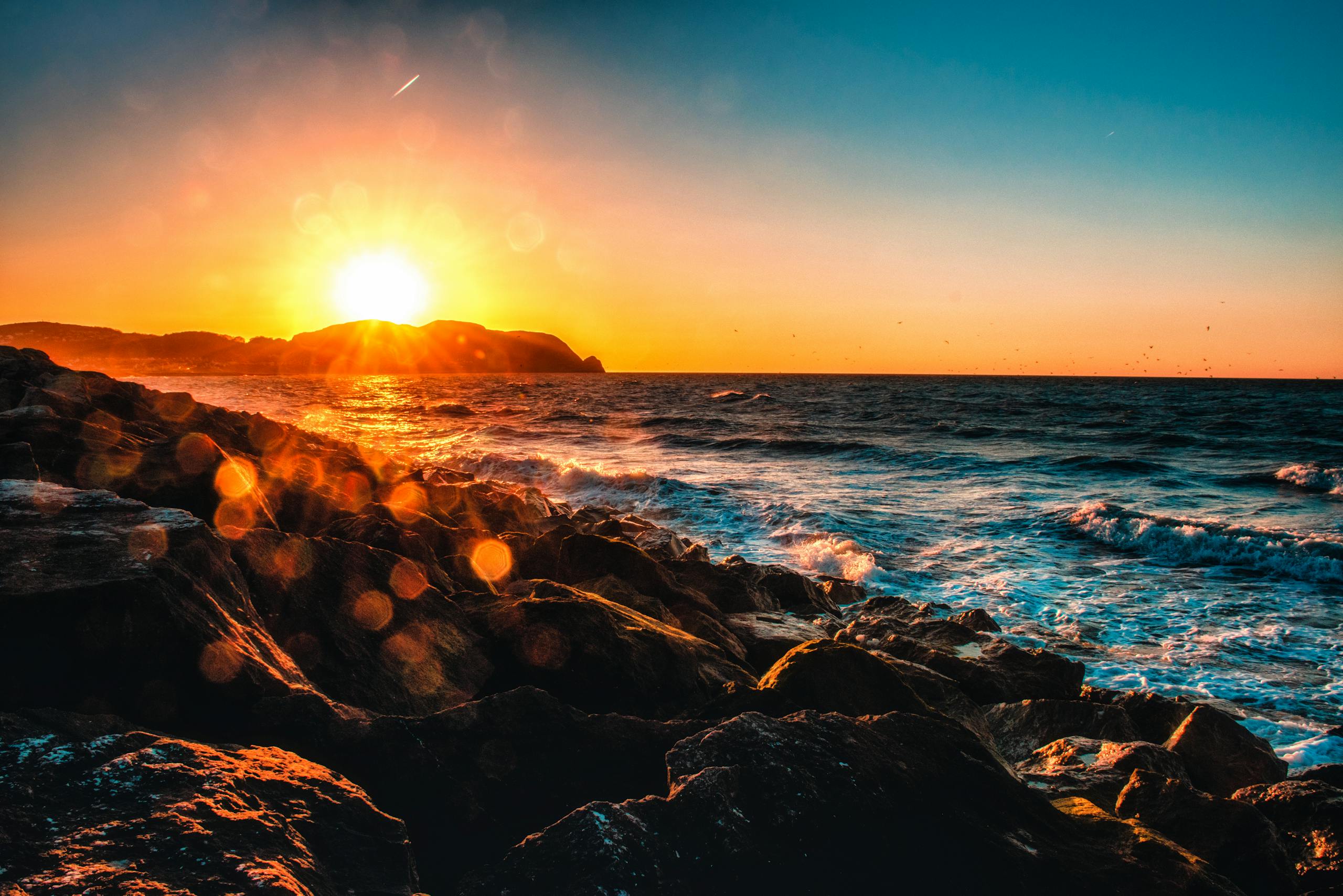 Captivating sunset view over a rocky seashore with vibrant sun glare and waves crashing.
